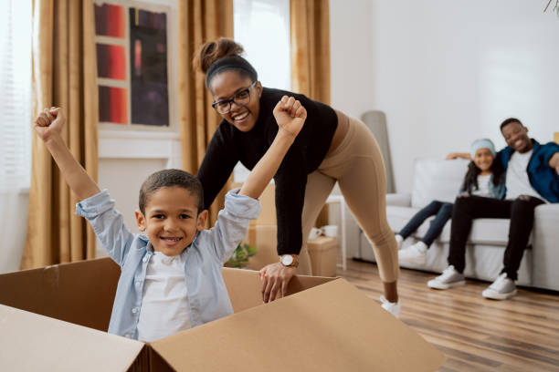 caring mom playing with baby, moving cardboard box across living room floor with sweet son sitting on it, joy of moving into new apartment, smiling little boy putting hands up from good time - family large american culture fun imagens e fotografias de stock