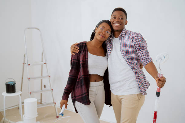 istock Handsome dark-skinned boy embraces beautiful smiling woman hold wall paint rollers in hand, couple hugs, together renovate new apartment, refresh white wall color 1377845215
