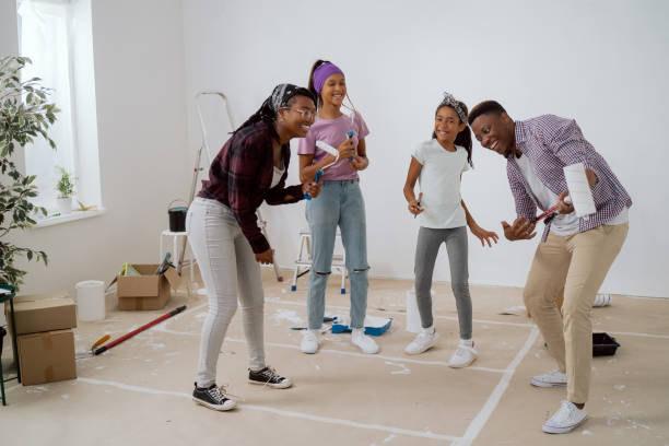 loca familia feliz baila en medio de la habitación durante el descanso de pintar las paredes, una mujer y sus hijas cantan a los rodillos, un hombre finge tocar la guitarra, divertida renovación de apartamento - home improvement couple painting real estate fotografías e imágenes de stock