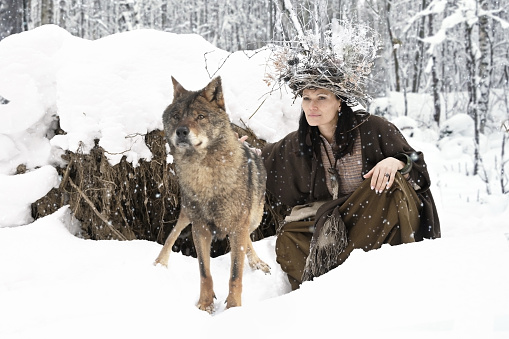 Female forest dweller with a hat made of twigs on her head and with a big gray wolf in the winter forest