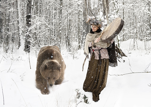 Shaman woman playing on shaman frame drum in a winter forest, next to her is a connecting rod bear