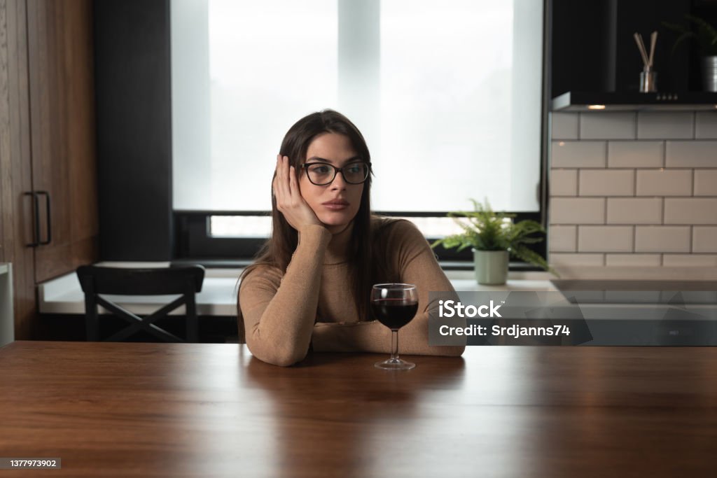 Young sad depressed woman sitting alone at home drinking wine feeling abandoned. Anxious female suffering from depression after relationship breakup drinking alcohol drink in kitchen try not to cry. Alcohol - Drink Stock Photo