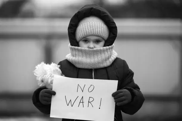 Little refugee girl with a sad look and a poster that says no to war. Social problem of refugees and internally displaced persons.