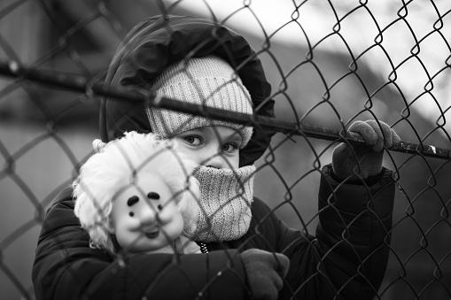 Little refugee girl with a toy behind a metal fence. Social problem of refugees and internally displaced persons.