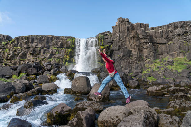 Mature women jumping on rocks against Oxararfoss waterfall at Mid-Atlantic Ridge, blue sky on sunny day, Iceland