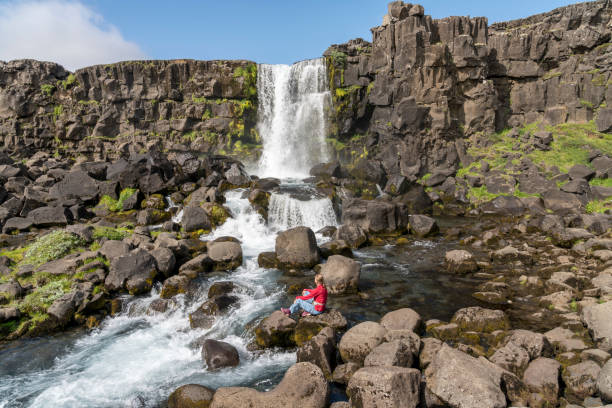 Mature women sitting on rocks against Oxararfoss waterfall at Mid-Atlantic Ridge, blue sky on sunny day, Iceland