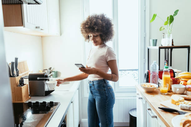Mixed race woman using smart phone in kitchen and preparing coffee from coffee machine African American woman working from home and drinking coffee Iphone stock pictures, royalty-free photos & images