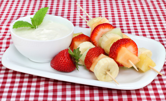 Skewers of fresh fruit kabobs with a side of yogurt dip, garnished with mint leaves.