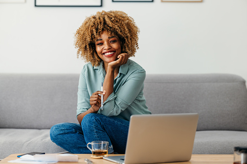 Smiling Afro-American woman is sitting on a sofa while looking at a camera. She might be working from home or studying. She is wearing a green shirt and jeans. She might be taking a break. There is a laptop and a cup of tea on the table in front of her.