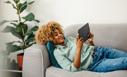 Cheerful Afro-American woman is lying on the sofa and watching something on a digital tablet. She is wearing a green shirt and jeans. She might be taking a break at work and enjoying some funny video or having a video chat with a friend. She might be a student.