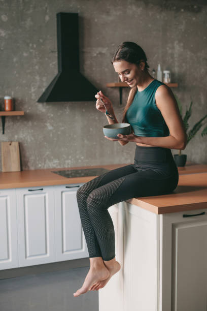 donna caucasica in abbigliamento sportivo che riposa in cucina dopo l'esercizio e mangia una ciotola di porridge. cibo sano vegetariano. - eating women breakfast cereal foto e immagini stock