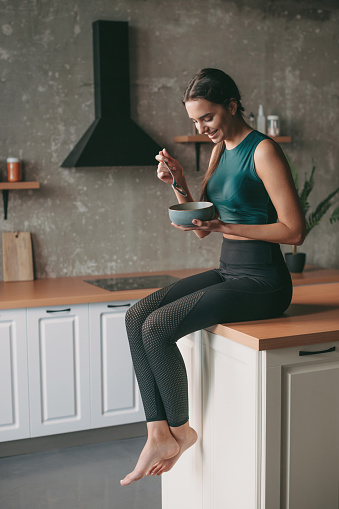 Caucasian woman in sportswear resting at the kitchen after exercise and eating a bowl of porridge. Vegetarian healthy food.