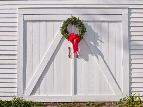 Green Christmas wreath with a red bow, hanging on white double barn doors.  Taken in bright sunlight.