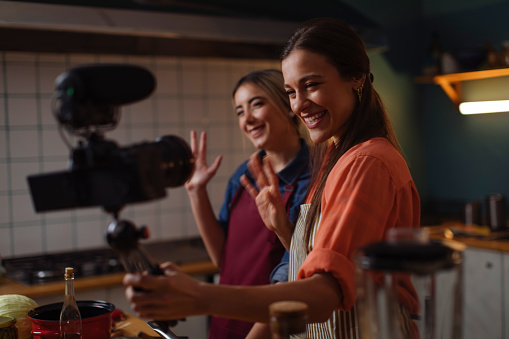 Two smiling women filming a cooking vlog while preparing food in the kitchen