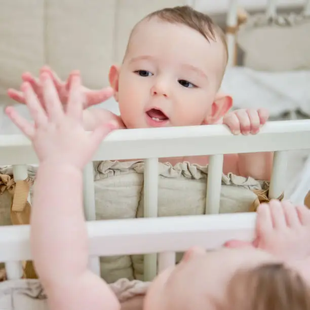 Happy baby is playing with her reflection in the mirror while standing in the crib. Funny child pulls his hand to the mirror, six months old