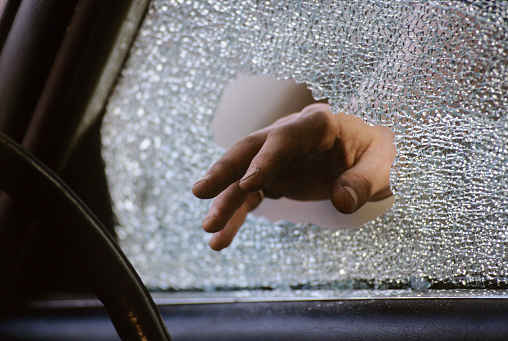 The hand of a thief (in semi silhouette) reaches in through a broken car windshield window