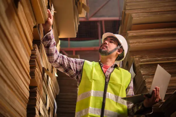 Asian male worker in safety uniform and hard hat, supervisor inspector with laptop for packaging stock order at factory warehouse, piles of stacking paper manufacture, recycling production industry.