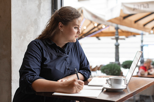 A smiling plus size woman working from a coffee shop, using her laptop and writing notes in a notebook.