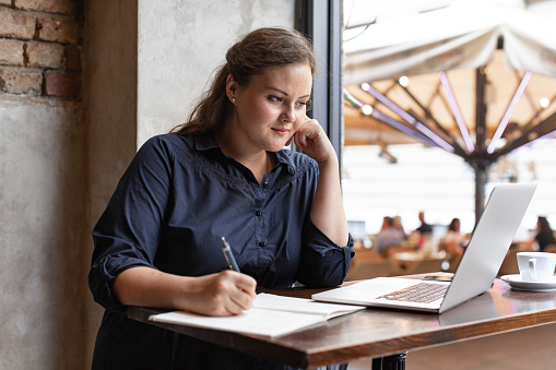 A smiling plus size woman working from a coffee shop, using her laptop and writing notes in a notebook.