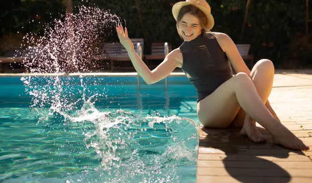 Photo of young beautiful caucasian woman sits in the summer on the shore of the pool in a straw hat and makes splashes of water with her hand.