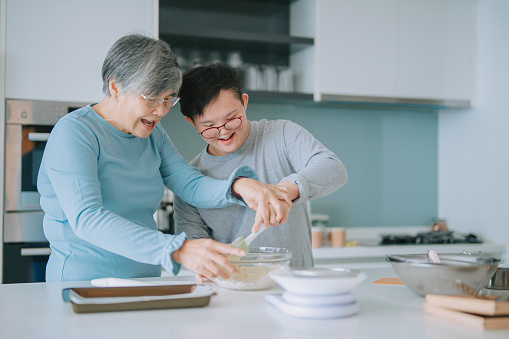 Asian Chinese senior woman teaching her disability down syndrome grandson preparing cake in kitchen mixing ingredient in bowl