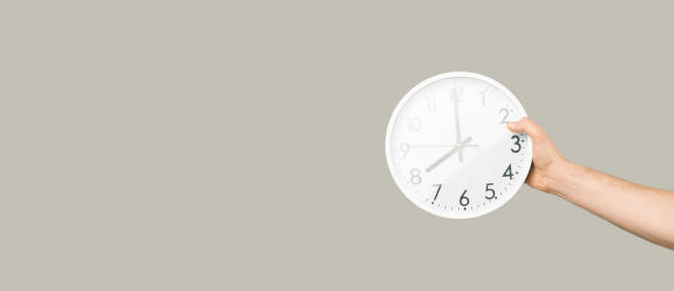 istock A male hand is holding a white wall clock over panorama grey background. 1378592639