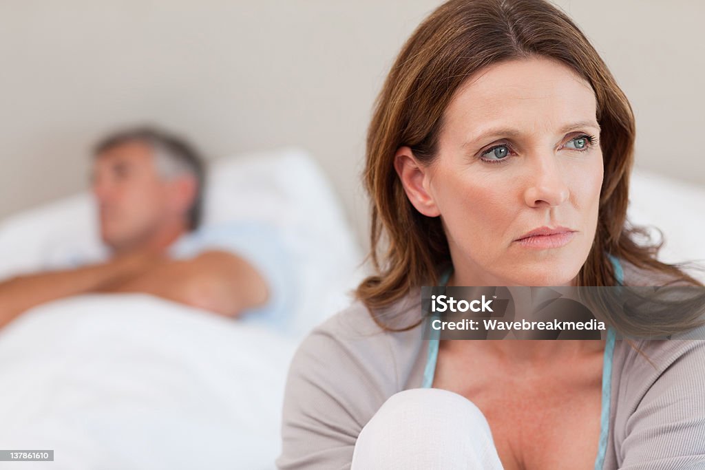 Sad woman on bed with her husband in the background Sad mature woman on bed with her husband in the background Mature Couple Stock Photo