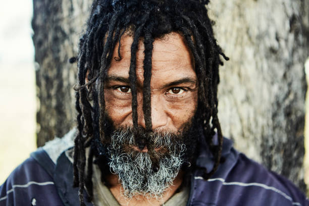 istock Homeless man with beard and dreadlocks makes strong eye contact with the camera as he stands against a tree 1378637532