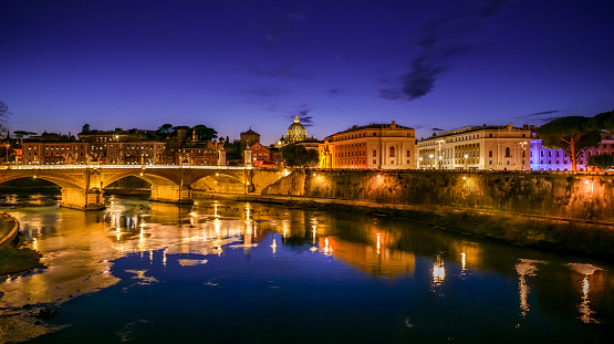 A suggestive and idyllic twilight view of the Tiber riverbank, in the historic heart of Rome. On the left, the Vittorio Emanuele II bridge, while on the horizon stands the imposing silhouette of the dome of St. Peter's Basilica. In 1980 the historic center of Rome was declared a World Heritage Site by Unesco. Super wide angle image in 16:9 and high definition format.