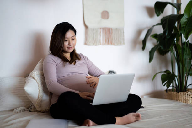 uśmiechnięta młoda azjatka w ciąży pracująca w domu, siedząca na łóżku z laptopa. styl życia w ciąży. ciężarna bizneswoman w pracy. pracująca matka w biznesie. home office, elastyczna koncepcja stylu życia pracy - aspirations occupation mother currency zdjęcia i obrazy z banku zdjęć