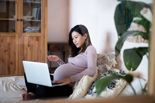 Professional Asian pregnant businesswoman working from home, having video call online meeting with business clients on laptop at home. Pregnancy lifestyle. Pregnant businesswoman at work. Working mother in business. Home office, flexible working lifestyle