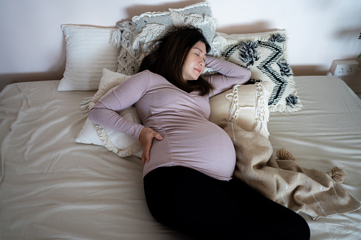 High angle shot of Asian pregnant woman holding her belly, sleeping on bed. Comfortable sleeping positions during pregnancy. Insomnia, sleeping disorder and physical discomfort during pregnancy