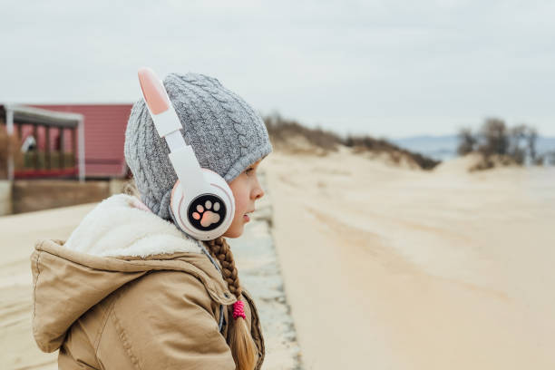 mała dziewczynka słuchająca muzyki w różowych słuchawkach dla dzieci z kocimi uszami na plaży jesienią, wiosną nad brzegiem morza poza sezonem, w sezonie nieturystycznym - six animals audio zdjęcia i obrazy z banku zdjęć