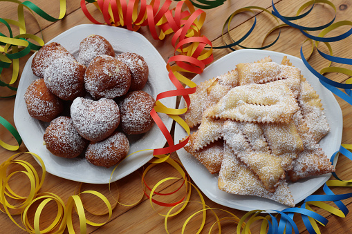 Traditional italian Crostoli ( also called  chiacchere, frappe, bugie, cenci, galani) and fritters called Frittelle with cream on wooden table with Confetti and coloful Carnival decorations