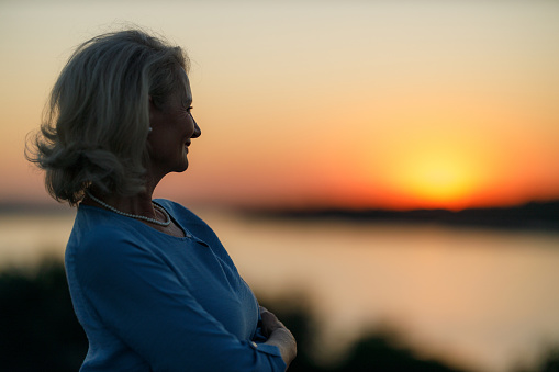 Smiling senior woman day dreaming while standing in nature with crossed arms at sunset. Copy space.