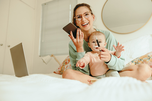 Happy mom taking a phone call while sitting on a bed with her baby. Cheerful single mother making plans with her clients while working from home. New mom balancing motherhood and work.