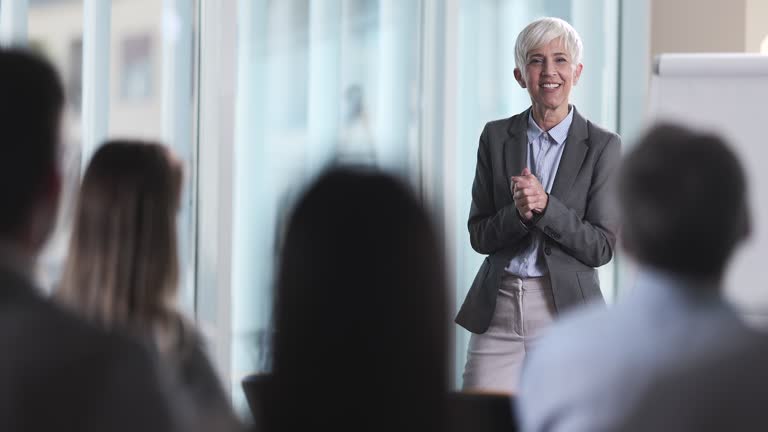Happy senior business leader giving a successful speech to large group of her colleagues who are applauding her in a board room.