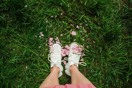 woman legs in white sneakers on green grass with pink blooming sakura flowers copy space