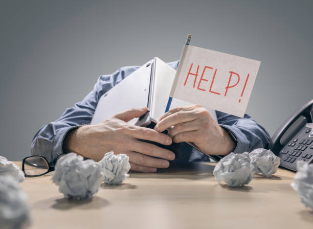 frustrated and overworked businessman burying his head under a laptop computer asking for help - crumpled paper document frustration imagens e fotografias de stock