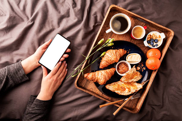istock Woman using smartphone while eating continental breakfast in bed. Blank screen copy space. 1378885427