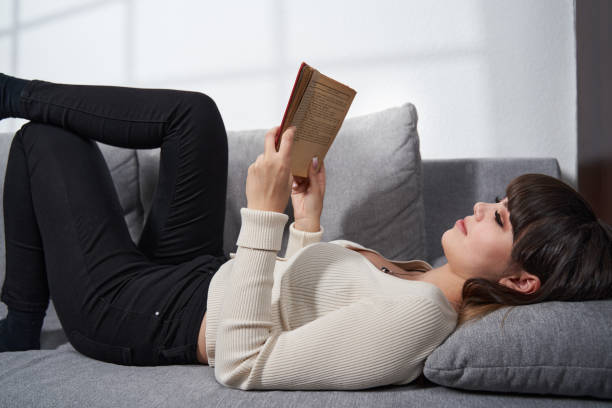 istock Young woman reading a book, sitting on sofa 1378897768
