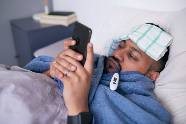 istock A sick man in bed with a thermometer looks at his mobile device 1378909507