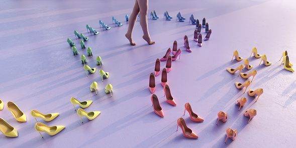 A woman with bare feet and legs standing on tiptoes in the middle of a looping line of high heel shoes arranged on the floor. The shoes are ordered in rainbow colours which gradually change in hue along the length of the line.