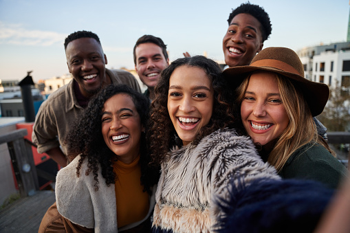 Multi-cultural group of friends taking a selfie at rooftop party. Close up, smiles with diverse young adults.
