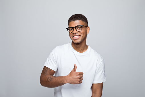 Happy african young man wearing white t-shirt and eyeglasses, standing with thumb up and laughing at camera. Studio portrait on grey background.