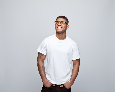 Happy african young man wearing white t-shirt and eyeglasses, standing with hands in pockets, looking away and smiling. Studio portrait on grey background.