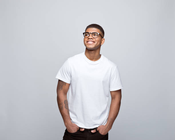 Portrait of friendly young man Happy african young man wearing white t-shirt and eyeglasses, standing with hands in pockets, looking away and smiling. Studio portrait on grey background. t shirt stock pictures, royalty-free photos & images