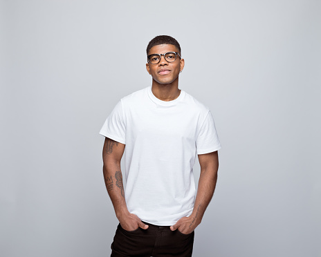Happy african young man wearing white t-shirt and eyeglasses, standing with hands in pockets and smiling at camera. Studio portrait on grey background.
