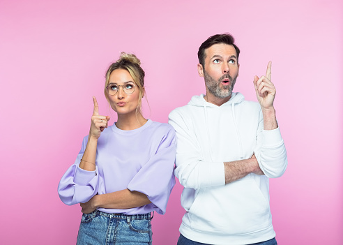 Mid adult couple pointing while looking up against pink colored background.