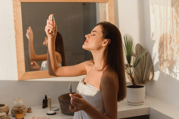 A beautiful young woman, wrapped in a towel, sprays mineral water on her face in front of a mirror. Skin hydration concept. A beautiful young woman, wrapped in a towel, sprays mineral water on her face in front of a mirror. Skin hydration concept heat haze stock pictures, royalty-free photos & images